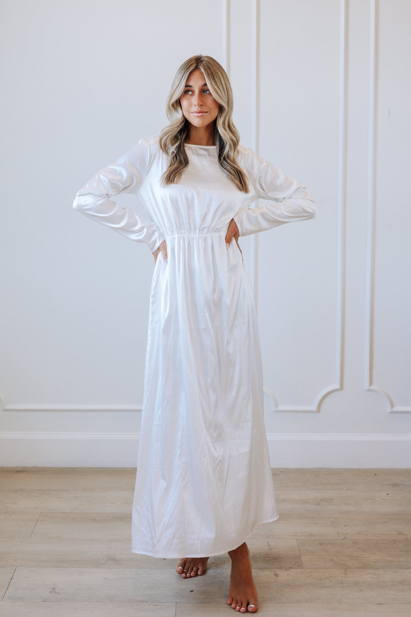 Woman wearing a long white dress standing against a white wall.