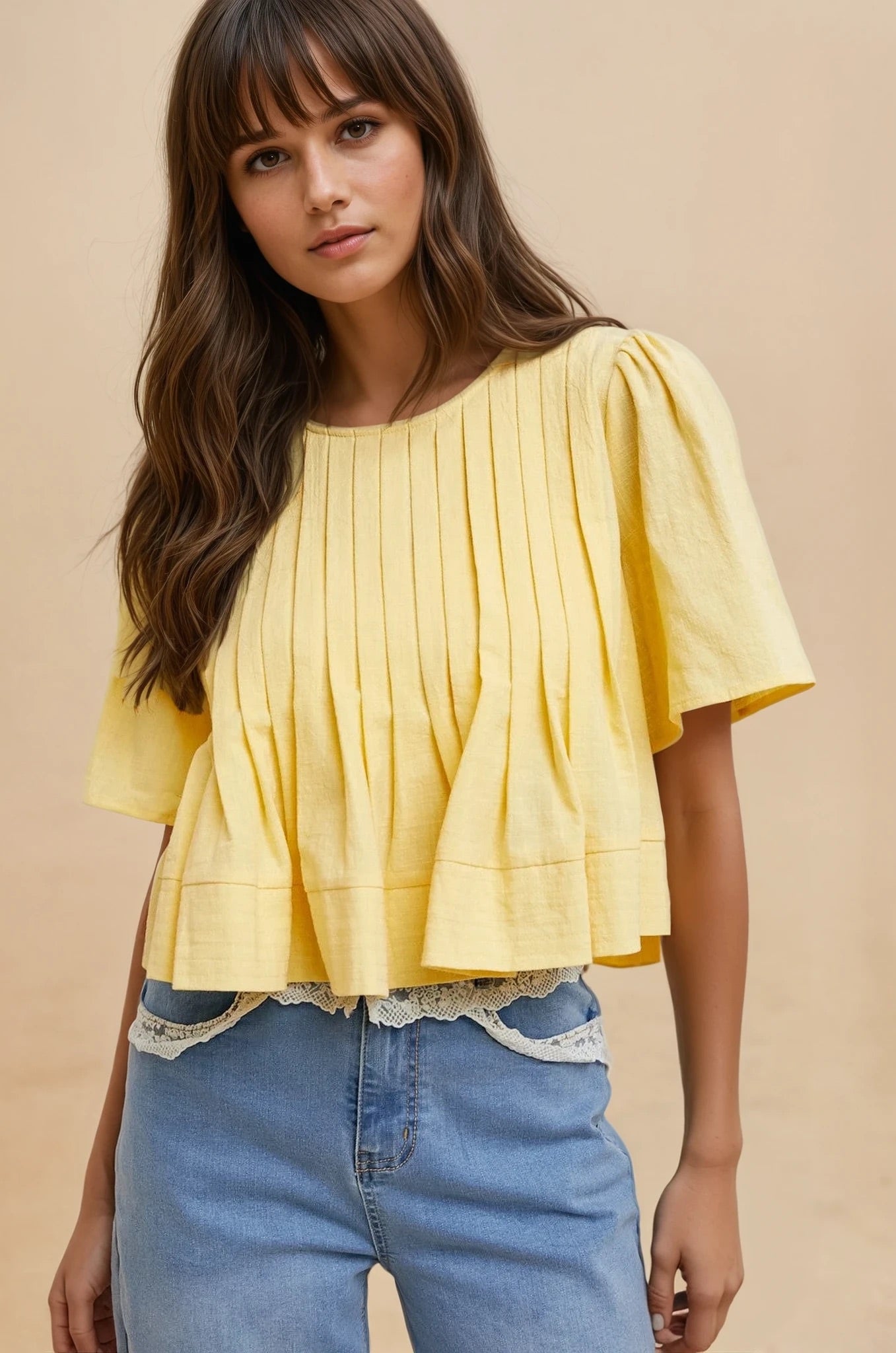 Woman wearing a yellow pleated top and blue jeans against a beige background
