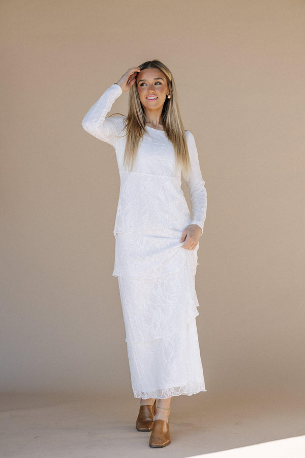 A model wearing a white maxi dress with lace details and a round neckline, standing against a beige background.