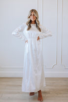 Woman wearing a long white dress standing against a white wall.