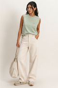 Woman wearing a light green sleeveless top and white pants, holding a beige bag against a plain background